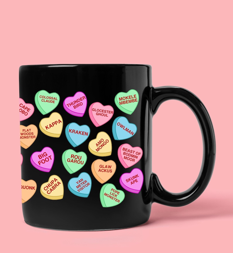 May include: Black ceramic mug decorated with colorful heart-shaped candies. Each candy has a different cryptid name, including "Bigfoot," "Kraken," and "Chupa Cabra." The mug has a curved handle and a pink background.
