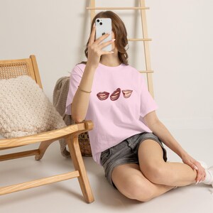 Puede incluir: Una camiseta de manga corta de color rosa claro con un estampado gráfico de tres pares de labios distintos en tonos marrones y rojos en el pecho. La camiseta tiene cuello redondo y un corte relajado, combinada con pantalones cortos de mezclilla gris oscuro.