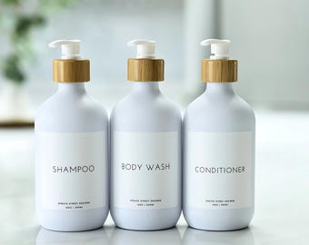 May include: Three white bottles with bamboo lids and pumps. The bottles are labeled "Shampoo", "Body Wash", and "Conditioner".