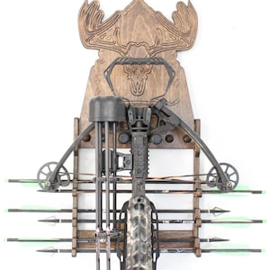 May include: A wooden crossbow wall mount with a carved moose head design. The mount holds a black crossbow and several arrows with green fletching. The background is white.