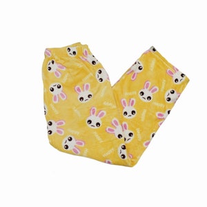 Puede incluir: Pantal&oacute;n de pijama amarillo con un patr&oacute;n repetido de conejos blancos con orejas rosas y la palabra "Happy" impresa en ellos.