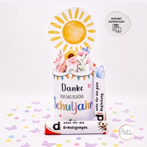 Puede incluir: Un envoltorio de regalo cilíndrico de papel con el texto Danke FÜR DAS SCHÖNE Schuljahr, decorado con flores en acuarela y un recorte de sol. Dos barras de chocolate están envueltas con el texto anke für die Begleitung y anke für die Ermutigungen.