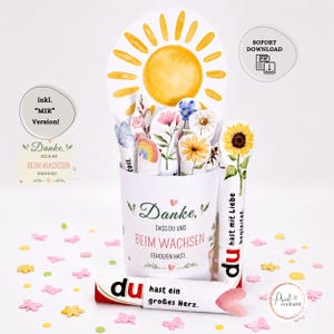 Puede incluir: Un recipiente cilíndrico blanco con el texto en alemán Danke, dass du uns beim Wachsen geholfen hast, lleno de recortes de papel de un sol amarillo, varias flores y un arcoíris. Dos tubos blancos con texto e ilustraciones florales están delante.