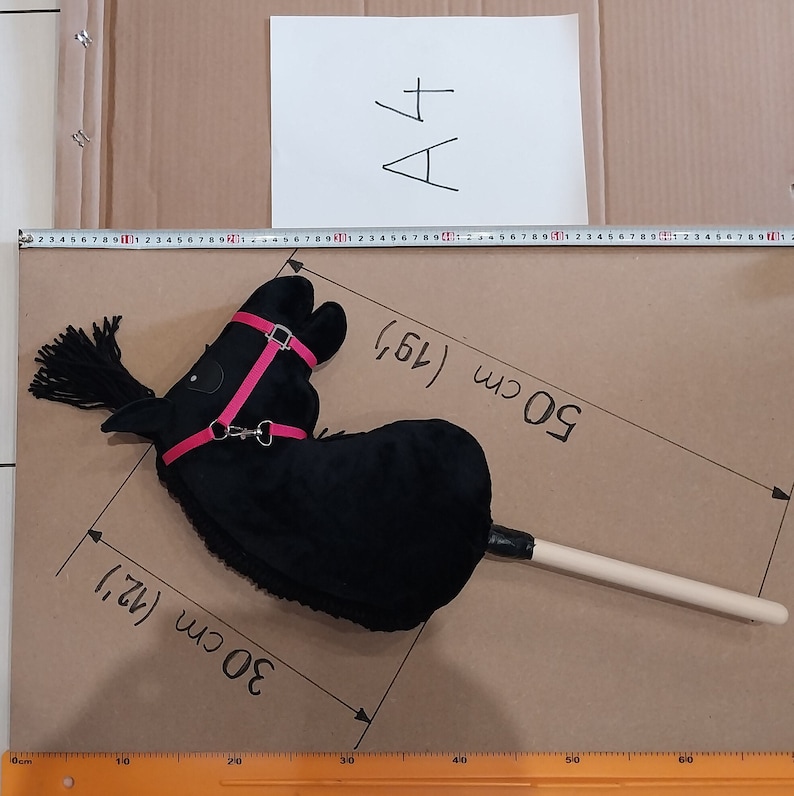 May include: A black plush hobby horse with a bright pink bridle and black yarn mane. The toy is attached to a wooden stick. A measuring tape and markings on the surface indicate a length of 50 cm and a width of 30 cm for the horse head.