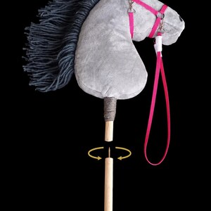 May include: A grey plush hobby horse with a dark grey yarn mane, featuring a bright pink bridle and a long pink lead. The horse head is attached to a detachable wooden stick handle, shown separated with a visible metal screw and yellow arrows.