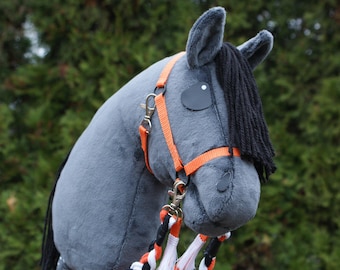 May include: A grey plush hobby horse on a wooden stick featuring a black yarn mane, black felt eyes, and an orange nylon halter. The horse is equipped with a braided rein in black, white, and orange, accented with small white tassels at the ends.
