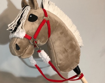 May include: A plush hobby horse with a tan fabric head, white and black yarn mane, and black fabric eyes. It features a red nylon bridle with silver-toned metal hardware and white faux fur trim on the noseband. The head is attached to a wooden stick.