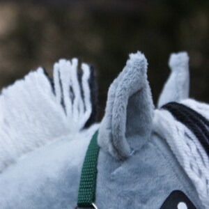 May include: A plush toy horse head made of soft gray fabric with a white and black yarn mane. The horse features a dark green woven nylon halter with a silver metal ring and a black textured fabric eye patch with a white circular pupil.