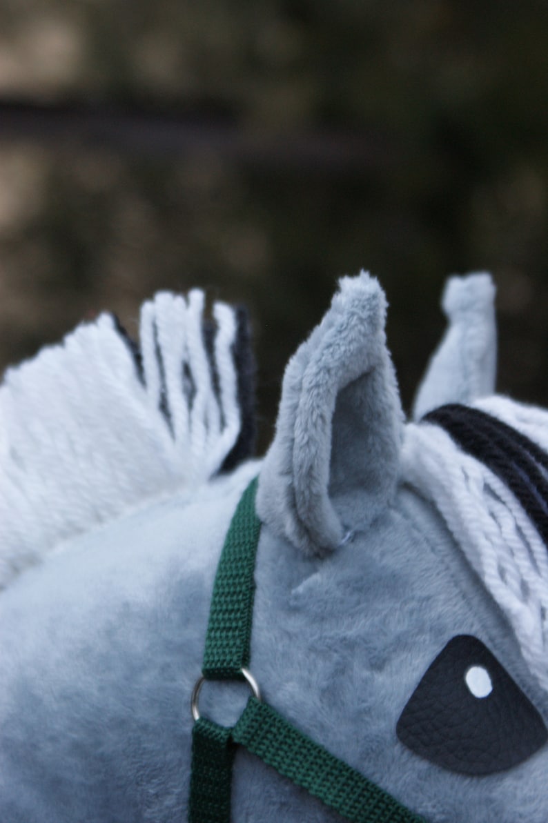 May include: A plush toy horse head made of soft gray fabric with a white and black yarn mane. The horse features a dark green woven nylon halter with a silver metal ring and a black textured fabric eye patch with a white circular pupil.