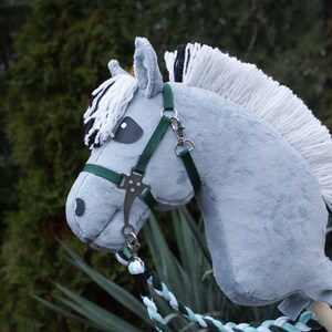 May include: A grey plush hobby horse with a white and black yarn mane and forelock. The horse features a dark green nylon bridle with silver-toned metal hardware and a braided lead rope in shades of white, mint green, and dark green.