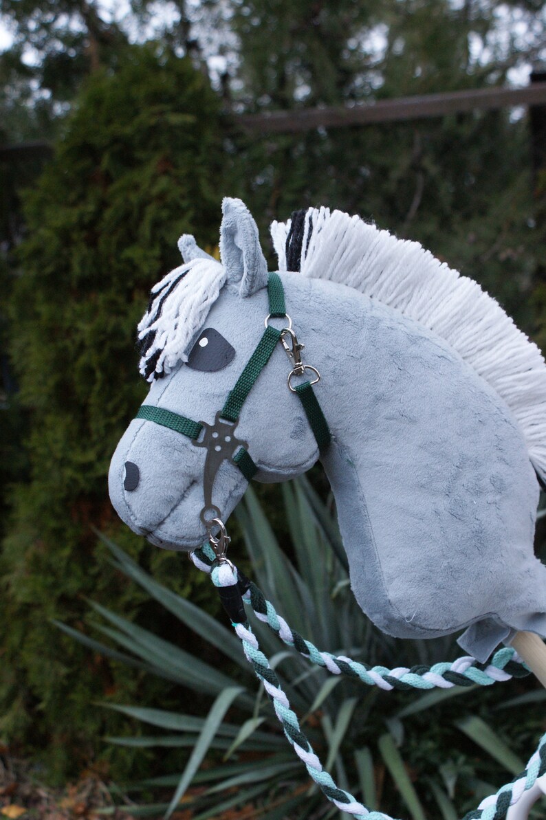 May include: A grey plush hobby horse with a white and black yarn mane and forelock. The horse features a dark green nylon bridle with silver-toned metal hardware and a braided lead rope in shades of white, mint green, and dark green.