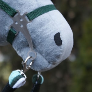 May include: A plush grey hobby horse head featuring a dark grey eye patch, a black nostril, and a white and black yarn mane. It is equipped with a dark green nylon bridle, a silver metal bit, and a braided lead rope in mint, white, and dark green.