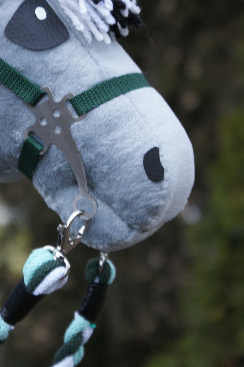 May include: A plush grey hobby horse head featuring a dark grey eye patch, a black nostril, and a white and black yarn mane. It is equipped with a dark green nylon bridle, a silver metal bit, and a braided lead rope in mint, white, and dark green.