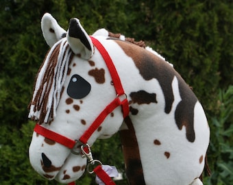 May include: A hobby horse toy featuring a plush white head with brown patches, a brown and white yarn mane, and black fabric eyes. The horse is equipped with a bright red nylon bridle and reins, and it is mounted on a light-colored wooden stick.