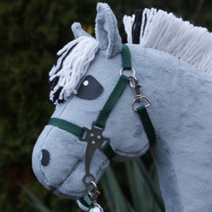May include: A grey plush hobby horse with a soft fabric texture, featuring a white and black yarn mane and forelock. It wears a dark green nylon halter with silver-toned metal hardware, including a bit and clips, attached to a braided lead rope.