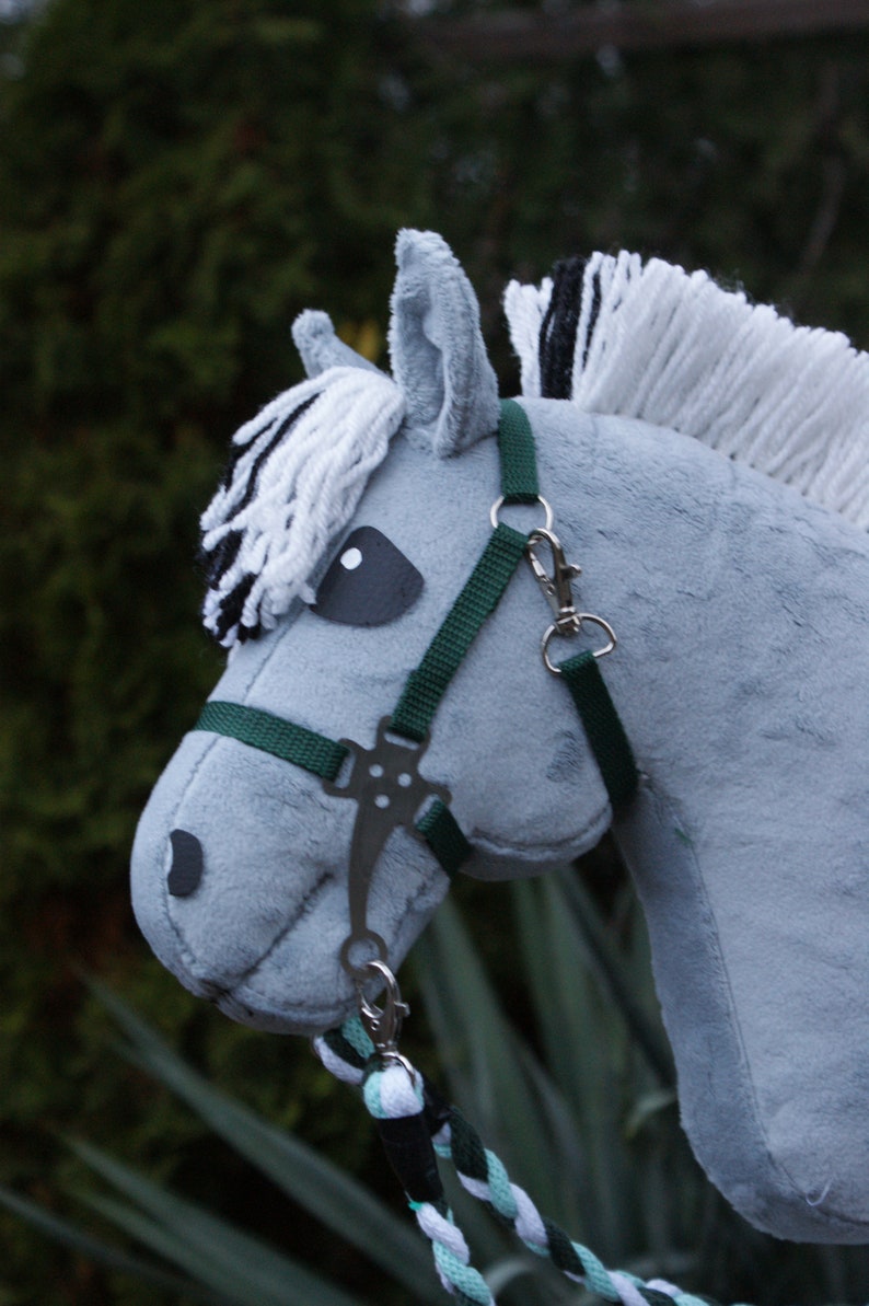 May include: A grey plush hobby horse with a soft fabric texture, featuring a white and black yarn mane and forelock. It wears a dark green nylon halter with silver-toned metal hardware, including a bit and clips, attached to a braided lead rope.