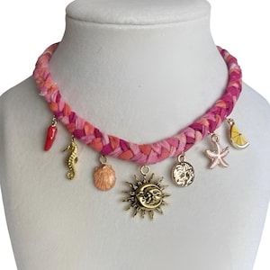 May include: A pink braided fabric necklace with various charms. Charms include a red chili pepper, seahorse, seashell, sun, coin, starfish, and lemon slice. The necklace is displayed on a white mannequin.