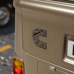 Könnte beinhalten: Ein schwarz-weißer Cummins-Logo-Aufkleber mit einem verzierten floralen Design ist auf der Rückseite eines beigefarbenen Fahrzeugs angebracht. Der Aufkleber hat die Form des Buchstabens "C" und enthält den Markennamen.