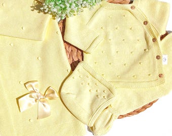 May include: A pale yellow knitted set consisting of a long-sleeved wrap-style top with small wooden buttons, a matching bonnet, and a blanket featuring a satin ribbon bow. The fabric has a raised dot texture throughout the entire garment set.