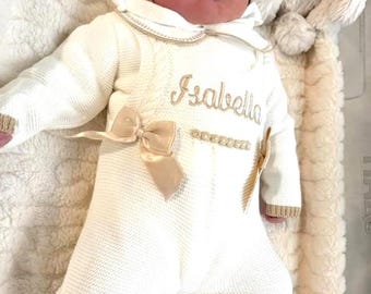 May include: A newborn baby wearing a cream-colored knitted romper with a matching hood. The romper features a bow, gold trim, and the name "Isabella" embroidered in gold thread. The baby is lying on a soft, cream-colored blanket.