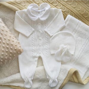 May include: A white knitted baby outfit set, including a romper with a Peter Pan collar, a hat, a blanket, and mittens. The romper features a cable knit design and button closures. The set is displayed on a soft, textured surface.