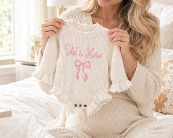 May include: A cream-colored long-sleeve knit baby bodysuit featuring a ruffled hem and cuffs. The front displays the embroidered pink text "She's Here" above a pink bow graphic. The garment includes three small brown buttons at the bottom.