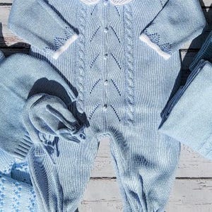 May include: A light blue knitted baby outfit set. The set includes a one-piece romper with a white collar, a matching hat, and a blanket. The romper features a cable knit design down the front and button closures. The outfit is made of soft, knitted material.