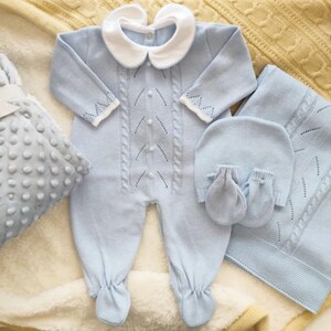 May include: A baby blue knitted romper with a white collar, matching hat, mittens, and blanket. The romper features a cable knit design with decorative stitching and button details. The set is displayed on a soft, textured surface.