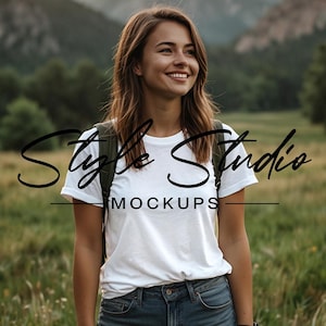 Op de afbeelding: Een vrouw draagt een wit T-shirt met de tekst "Style Studio Mockups" erop. Ze staat in een veld met groen gras en een rugzak.