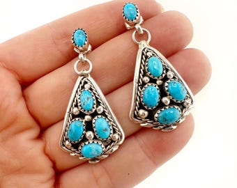 May include: A pair of sterling silver dangle earrings featuring oval-shaped turquoise stones. Each earring has a single turquoise stone at the top and a triangular base set with three additional turquoise stones, accented by twisted wire and silver beads.