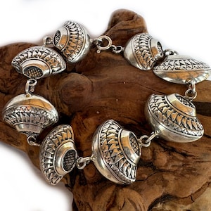 Puede incluir: Pulsera plateada con una serie de eslabones decorativos en forma de cúpula. Cada eslabón tiene un patrón intrincado y texturizado. La pulsera se muestra sobre un trozo de madera natural.