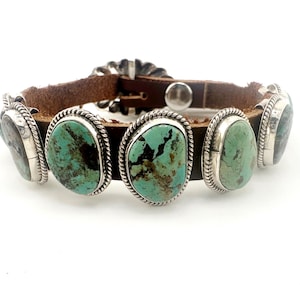 Pode incluir: Uma pulseira de couro castanho com cinco pedras turquesa ovais engastadas em prata com detalhes de corda torcida. A pulseira inclui um fecho de fivela de metal prateado e um detalhe decorativo de concho de prata no fecho.