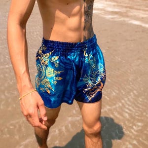 Op de afbeelding: Blauwe en gouden Muay Thai shorts met een traditioneel Thais ontwerp. De shorts zijn gemaakt van satijn en hebben een trekkoord.