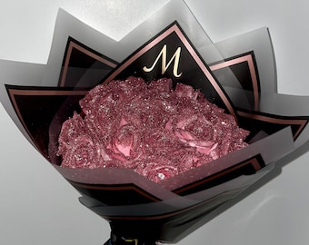 May include: A bouquet of pink glitter roses wrapped in black and gold paper with the letter "M" in gold lettering on the front.