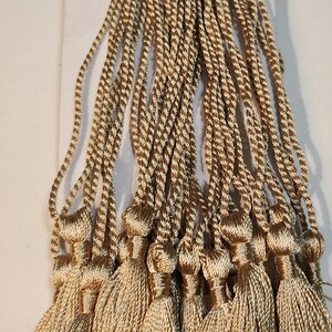 May include: A bundle of gold-colored tassels, each with a twisted cord and a full, fringed bottom. The tassels are held together with a red rubber band and are arranged on a white card.