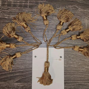 May include: A collection of ten brown tassel trim with a gold sheen. The tassels are attached to cords and are arranged in a fan shape.