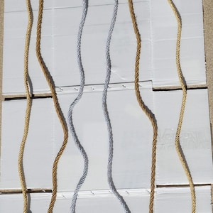 May include: Three decorative tassels with gold and silver cords. The tassels are hanging from a white surface. The tassels are made of a braided cord with a tassel at the end. The tassels are different lengths.