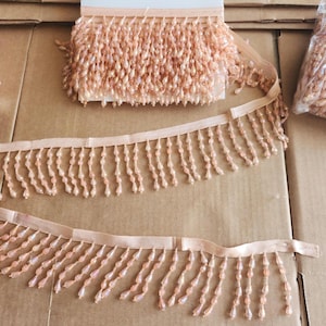 May include: Peach-colored beaded fringe trim. The trim features rows of small, teardrop-shaped beads hanging from a sheer ribbon. A roll of the trim is visible, along with two lengths of the trim unrolled.