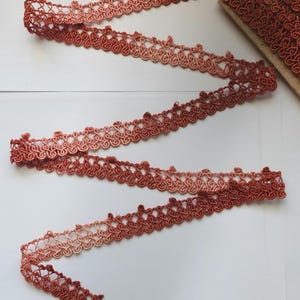 May include: A close-up of a length of ombre trim with a delicate, intricate design. The trim is a gradient of peach, coral, and rust colors.