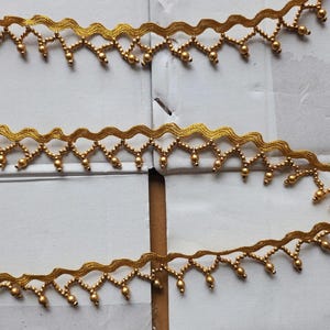 May include: Close-up of gold-colored decorative trim. The trim has a scalloped edge with small, dangling beads. The design features a repeating pattern of zigzags and curves, creating a delicate and ornate appearance. The trim is likely used for embellishing clothing or craft projects.