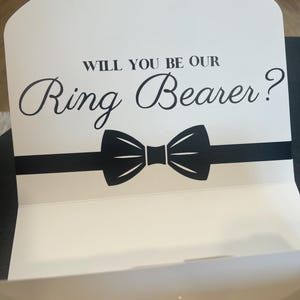May include: White card with black text and a black bow tie. The text reads "Will you be our Ring Bearer?"