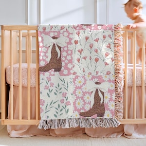 May include: A woven baby blanket with a patchwork design. The blanket features brown cowboy boots, white bows, and floral patterns in pink, white, and green. The blanket has a fringed edge and is draped over a wooden crib.