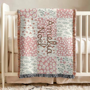 May include: A patchwork baby blanket with a floral pattern in pink and white, featuring the name "Amelia Noel" in brown script. The blanket has a fringed edge and is draped over a crib.
