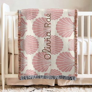 May include: A cream-coloured baby blanket featuring a pattern of pink seashells. The name "Olivia Rae" is printed vertically in brown script. The blanket, with a fringed edge, is draped over a white cot.