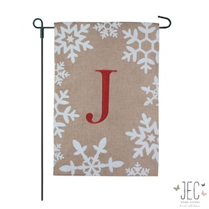 May include: A tan garden flag with white snowflakes and a large red letter "J". The flag is displayed on a black metal stand. The flag is a seasonal home decor item.