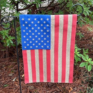 May include: A garden flag featuring the American flag design. The flag has a blue square with gold stars in the upper left corner and red and tan stripes. The flag is hanging on a black metal garden stake.