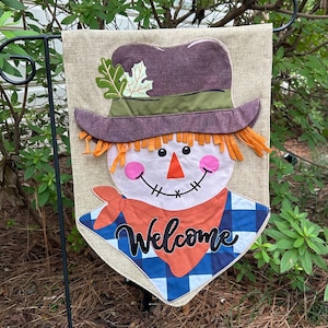 May include: A burlap garden flag featuring a scarecrow design. The scarecrow has a purple hat, orange hair, a pink nose, and a blue and white checkered shirt. The word "Welcome" is written in cursive on the orange bandana.