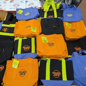 Könnte beinhalten: Verschiedene Trader Joe's Canvas-Tragetaschen in verschiedenen Farben, darunter Orange, Schwarz und Blau. Jede Tasche zeigt das Trader Joe's-Logo in einem kreisförmigen Design. Einige Taschen haben farblich abgesetzte Griffe.