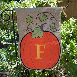 May include: A decorative garden flag featuring an orange pumpkin with a black outline and the letter "F" in gold. Green leaves and vines adorn the top of the pumpkin. The flag is made of a burlap-like material and is displayed on a black metal stand.