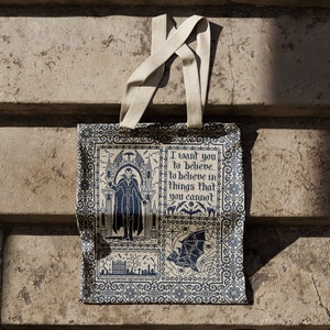 Puede incluir: Una bolsa de tela beige con un dise&ntilde;o estampado en azul y blanco. La bolsa presenta una ilustraci&oacute;n de estilo g&oacute;tico de un hombre con una capa y una cita que dice "Quiero que creas, que creas en cosas que no puedes."